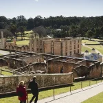 Two people walk down a paved path towards the ruins of a large historic brick building at the Port Arthur Historic Site, surrounded by green grass and trees under a clear sky near Hobart.