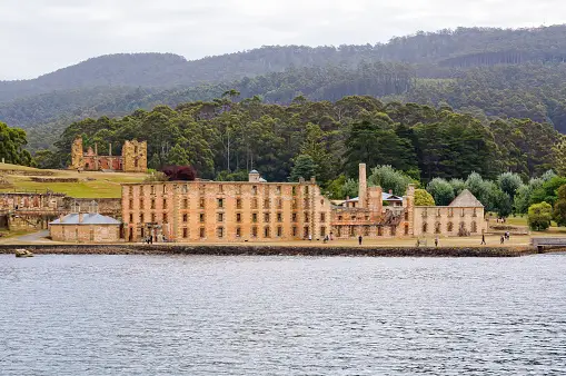 Historic stone buildings of the Port Arthur Historic Site stand by the water, with forested hills in the background under an overcast sky, offering a glimpse into Tasmania’s convict past just a short drive from Hobart.